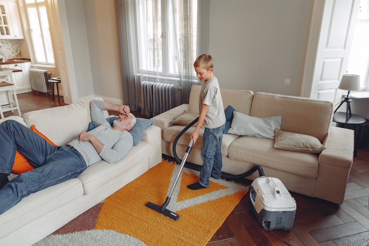 hero-img-02 A young boy vacuums the living room carpet while his grandfather relaxes on the sofa.